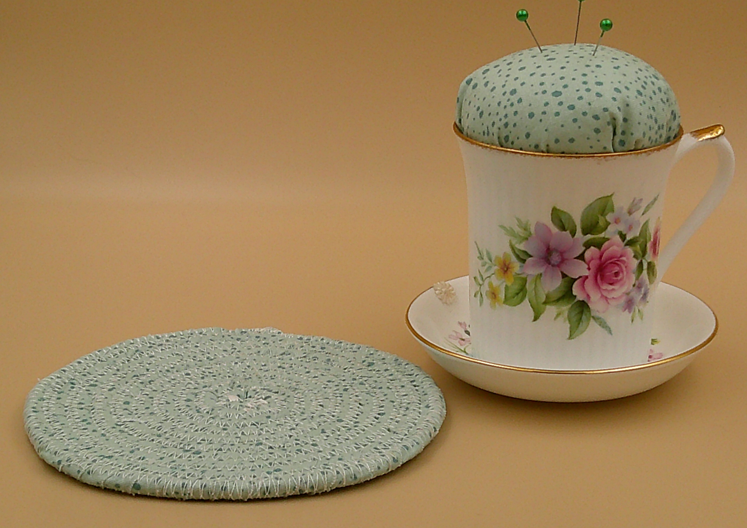 This shows the pincushion bobbin holder next to the trivet. The trivet is about 5.5 inches wide. Flower is on saucer to left of cup. This shows the pincushion bobbin holder next to the trivet. The trivet is about 5.5 inches wide. Flower is on saucer to left of cup.